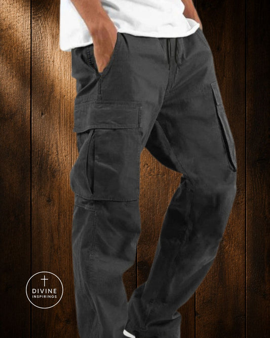 A person wearing dark charcoal cargo pants with a white t-shirt, standing against a dark wood plank background. A circular logo for 'Divine Inspiration' is visible in the bottom-left corner.