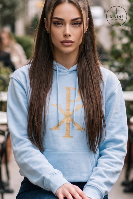 A woman wearing a light blue hoodie with a gold cross graphic on the chest, photographed in an outdoor setting. A circular 'DIVINE INSPIRINGS' logo is visible in the top right corner.