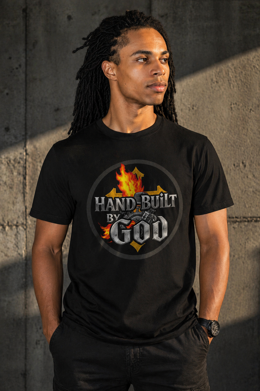 Man wearing a black t-shirt with 'Hand-Built by God' graphic design against a concrete wall.