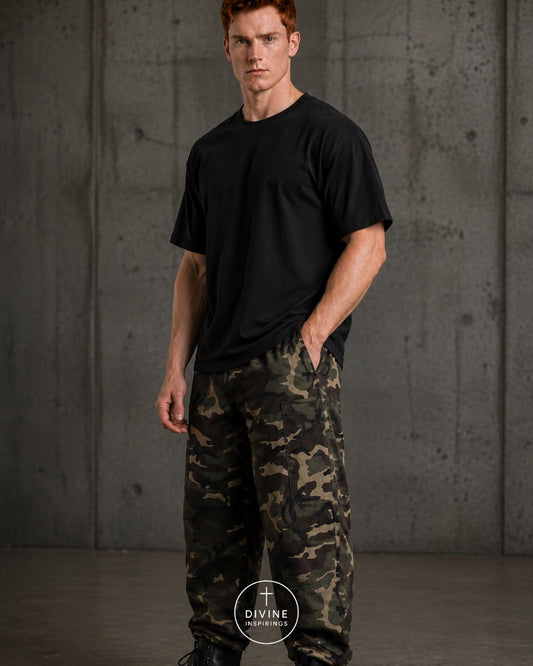 A man wearing a black crewneck t-shirt and camouflage cargo pants, standing against a textured gray concrete wall. A circular 'DIVINE INSPIRATIONS' logo is visible at the bottom center.