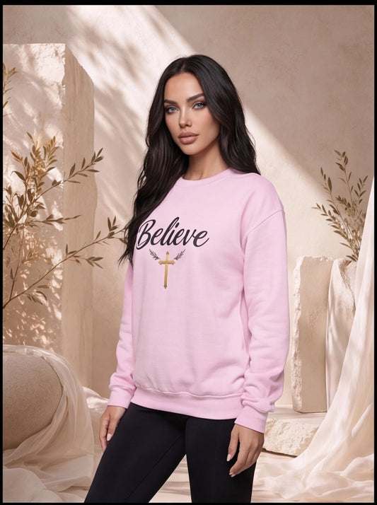 Woman wearing a pink sweatshirt with 'Believe' and a cross design, standing in a softly lit room.