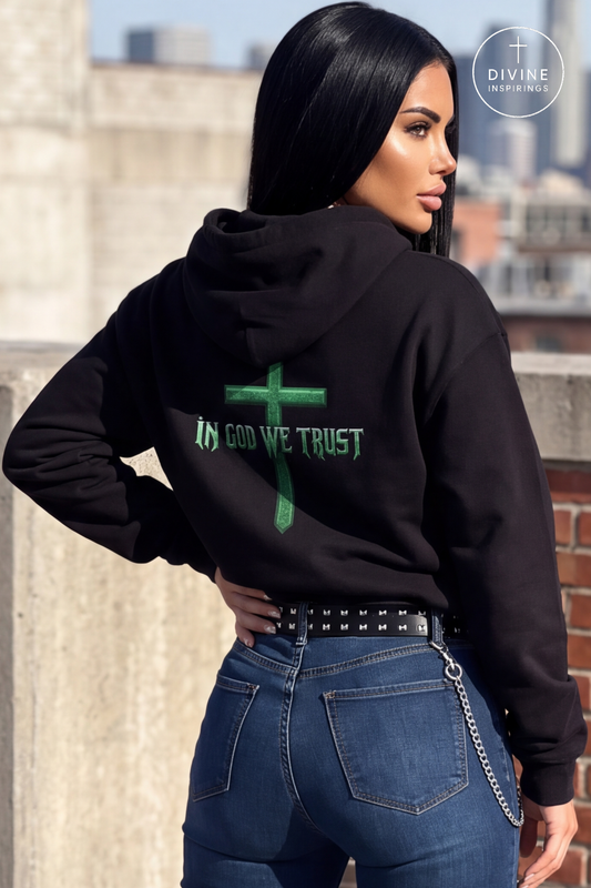 A woman wearing a black hoodie with a green cross and the text 'IN GOD WE TRUST' on the back, paired with blue jeans, photographed outdoors against a cityscape background.