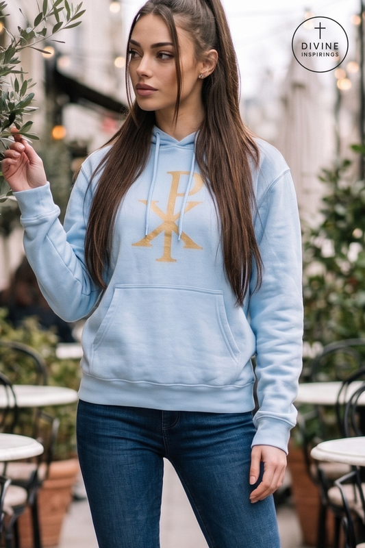 A woman wearing a light blue pullover hoodie with a gold Chi-Rho graphic on the chest, paired with dark blue jeans, standing in an outdoor cafe setting.