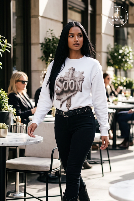 A woman walking outdoors on a city sidewalk wearing a white long-sleeve crewneck sweatshirt with a metallic 'SOON' graphic on the front, paired with black skinny jeans and a black studded belt. The background shows a blurred outdoor cafe setting with tables and chairs.