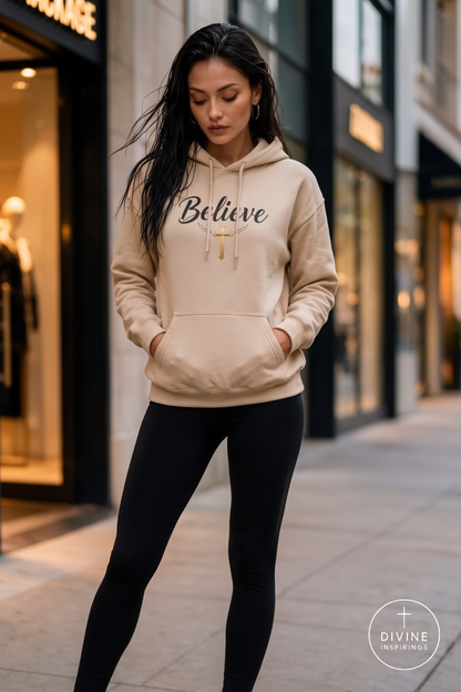 A woman wearing a beige hoodie with 'Believe' and a cross graphic printed on the chest, paired with black leggings, standing on a city sidewalk.