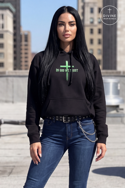 A woman with long black hair stands on a rooftop wearing a black pullover hoodie with a green cross and the text 'IN GOD WE TRUST' on the chest, paired with blue jeans and a black studded belt with a silver chain. A 'DIVINE INSPIRINGS' logo is visible in the top right corner.