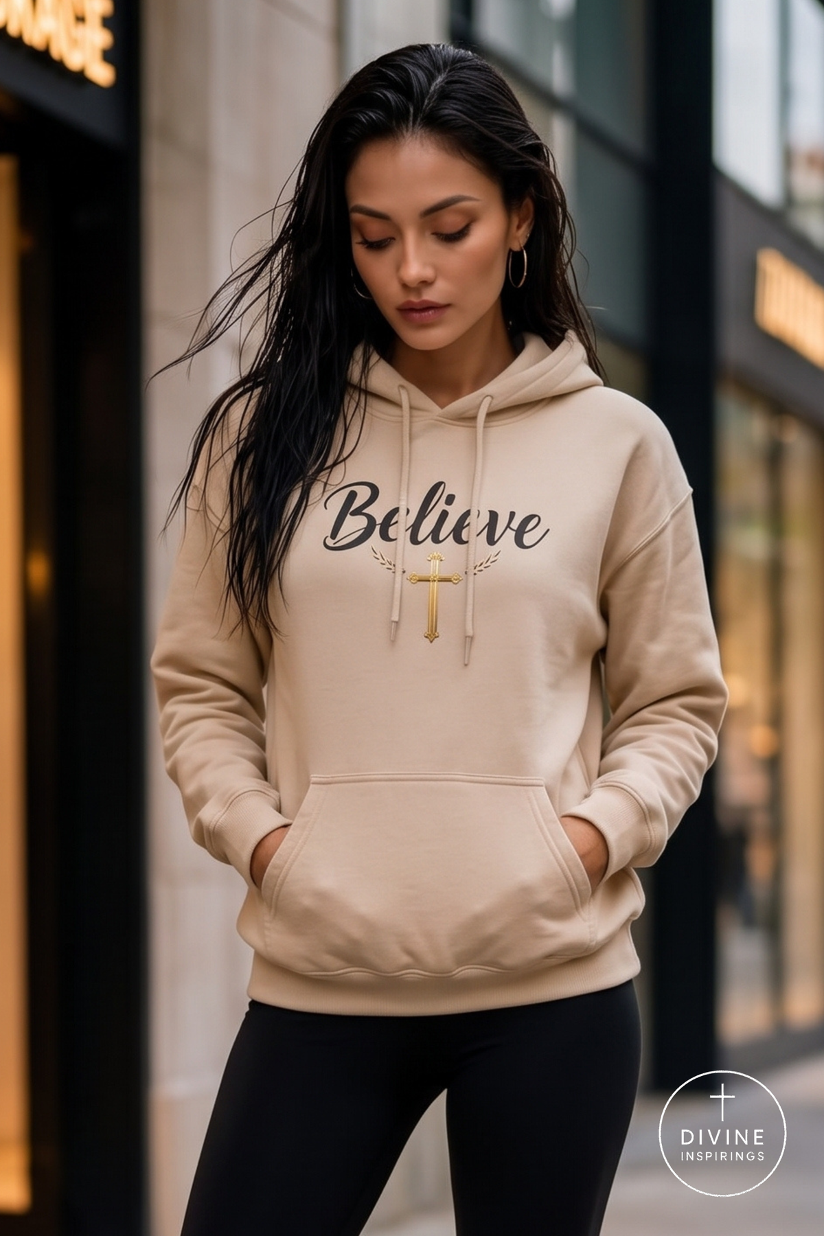 A woman wearing a beige hoodie with the word 'Believe' in black script and a gold cross graphic on the chest, standing outdoors in front of a blurred storefront.