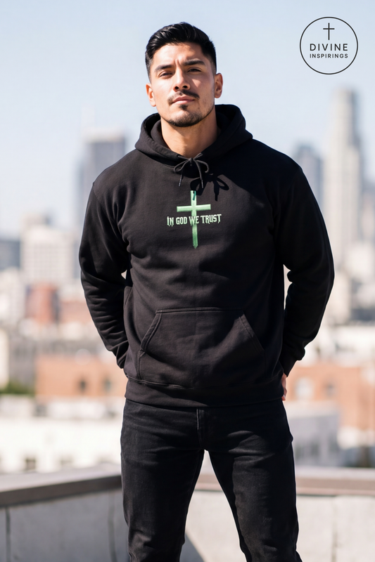 A man wearing a black pullover hoodie with a green cross and the text 'IN GOD WE TRUST' on the chest, standing on a rooftop with a blurred city skyline in the background. A circular 'DIVINE INSPIRINGS' logo is visible in the top right corner.