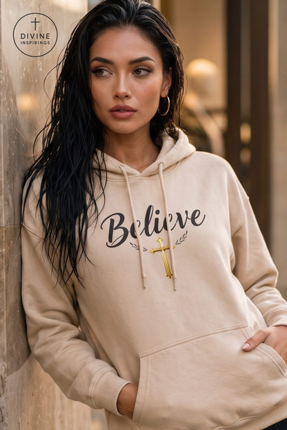 A woman wearing a beige hoodie with 'Believe' printed in black script and a small gold cross graphic, leaning against a stone wall. The Divine Inspirations logo is visible in the top left corner.