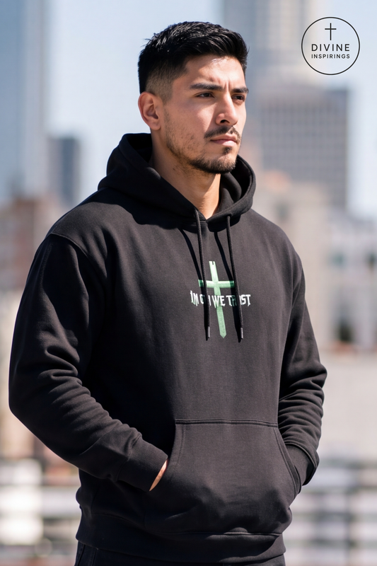 A man wearing a black pullover hoodie with a green cross and 'IN GOD WE TRUST' text on the chest, standing outdoors with a blurred city background. A circular 'DIVINE INSPIRINGS' logo is visible in the top right corner.