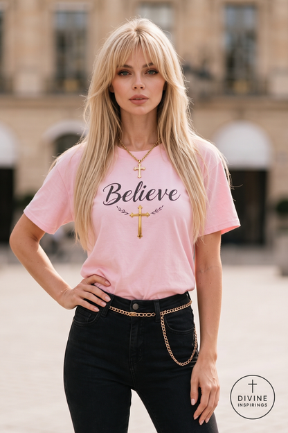 A blonde woman wearing a light pink t-shirt with 'Believe' and a gold cross printed on the front, paired with black jeans and a gold chain belt, standing in an outdoor urban setting.