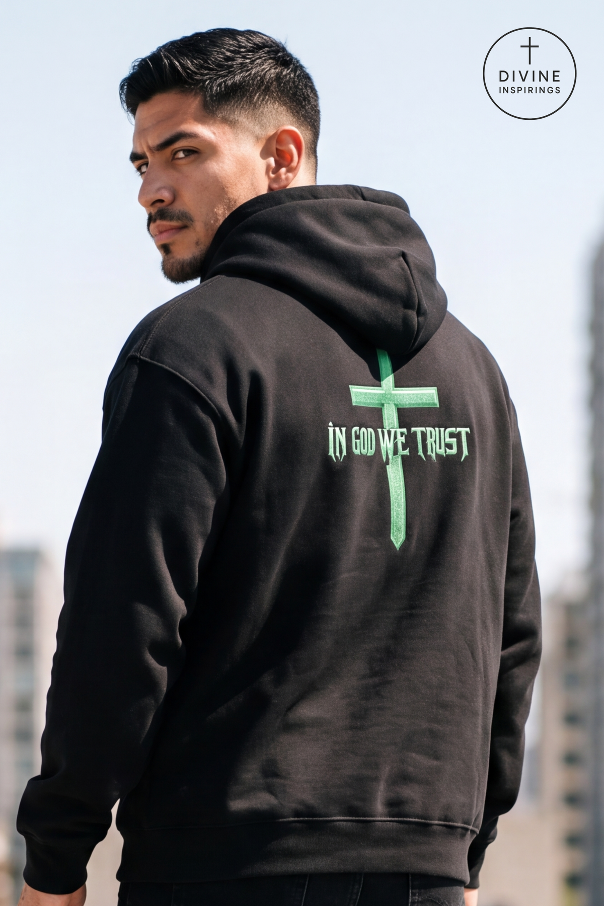 A man wearing a black hoodie with a green cross and the text 'IN GOD WE TRUST' printed on the back, shown from behind against an outdoor city background. A 'DIVINE INSPIRINGS' logo is visible in the top right corner.