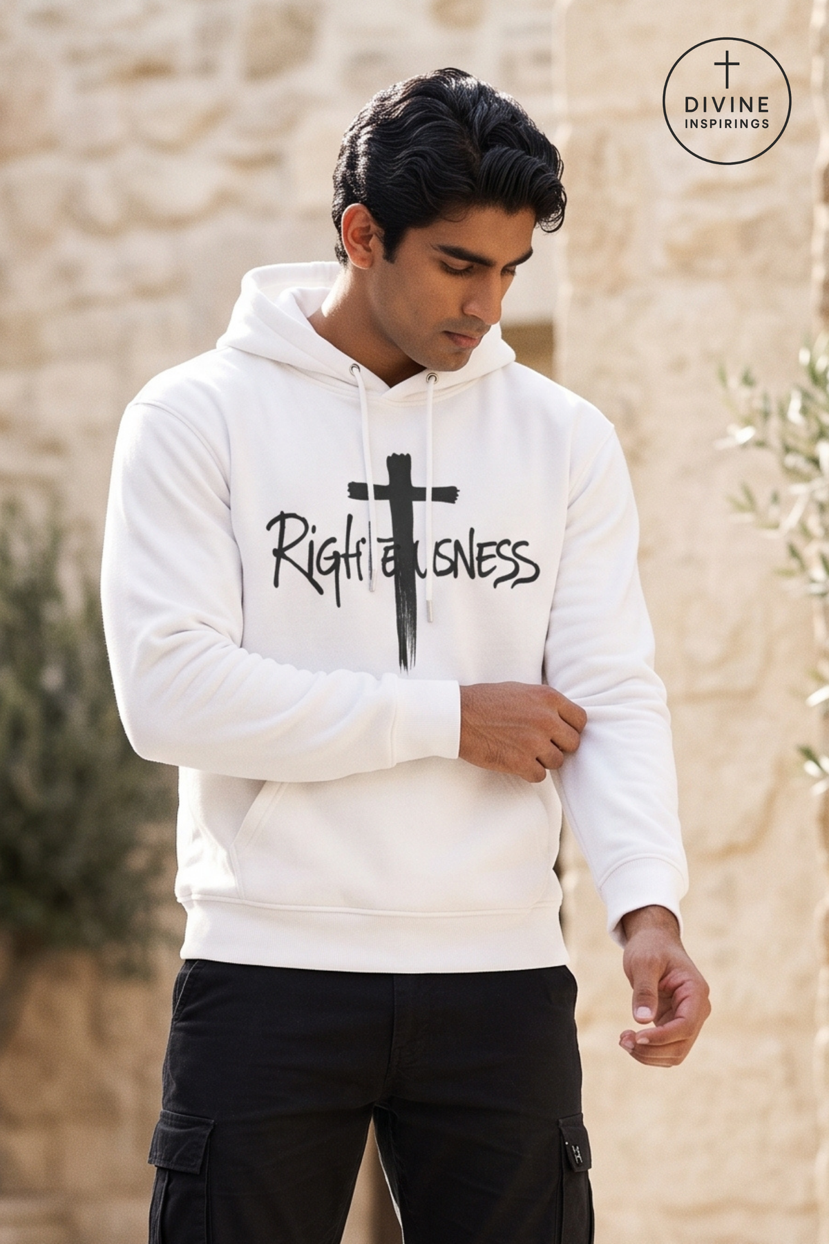 A man wearing a white pullover hoodie with a black cross graphic and the word 'Righteousness' printed on the front, paired with black cargo pants, standing outdoors against a beige stone wall. A circular 'DIVINE INSPIRINGS' logo is visible in the top right corner.
