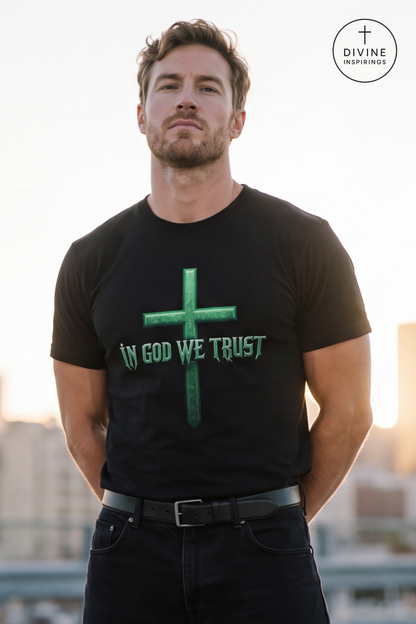 A man wearing a black t-shirt with a green cross and the text 'IN GOD WE TRUST' printed on the front, standing outdoors with a blurred city skyline in the background. A 'DIVINE INSPIRINGS' logo is visible in the top right corner.
