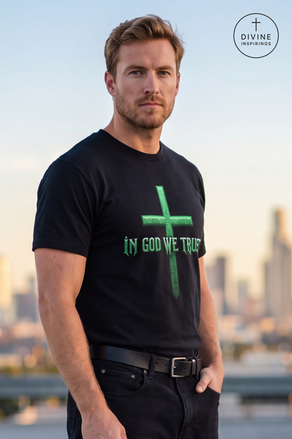 A man wearing a black t-shirt with a green cross and the text 'IN GOD WE TRUST' printed on the front, standing outdoors with a blurred city skyline in the background. A circular 'DIVINE INSPIRINGS' logo is visible in the top right corner.