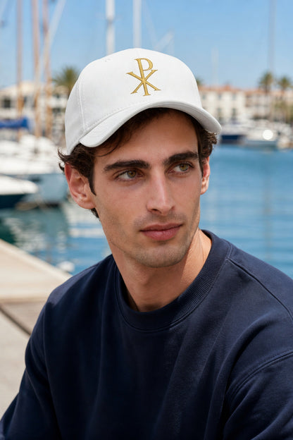 Man wearing a white cap with a logo, sitting by a waterfront with boats in the background.