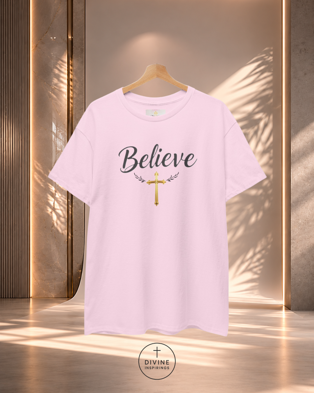 Pink t-shirt with 'Believe' and cross design on a hanger against a neutral background