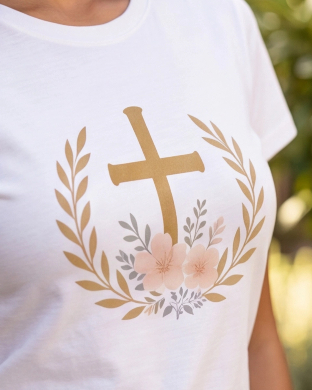 White t-shirt with a gold cross and floral design on a blurred natural background
