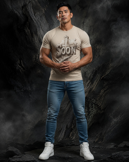 Man wearing a beige t-shirt with text and blue jeans standing against a dark rocky background