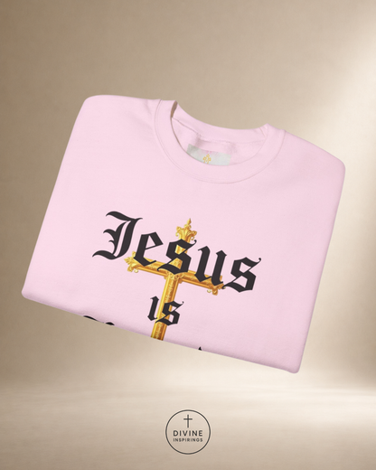 Folded pink sweatshirt with 'Jesus is' design on a beige background