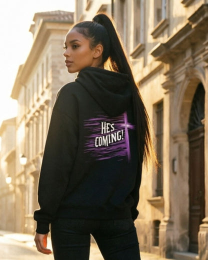 Person wearing a black hoodie with 'HE'S COMING!' text in a city setting