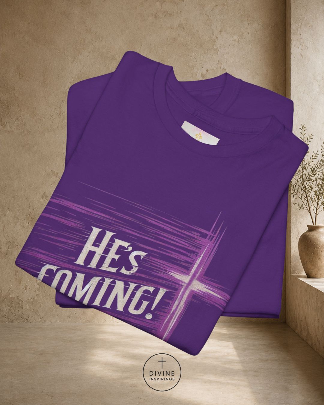 Folded purple t-shirt with 'HE'S COMING!' text on a beige surface.