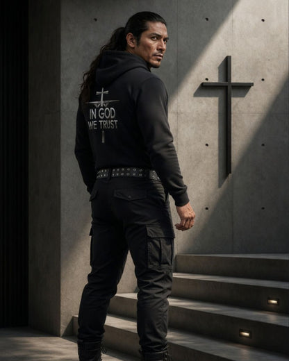 Man wearing a hoodie with 'In God We Trust' text, standing in a concrete setting with a cross.