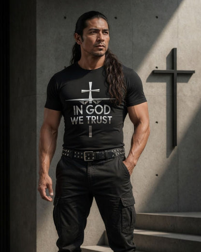 Man wearing a black t-shirt with 'In God We Trust' text, standing against a concrete wall with a cross.