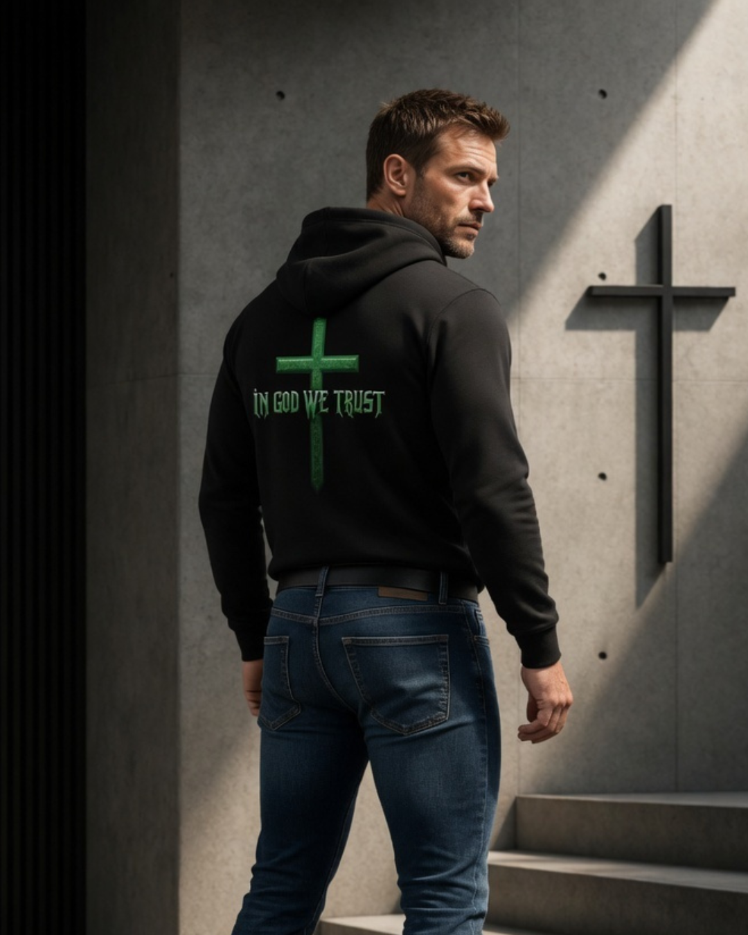 Man wearing a black hoodie with 'In God We Trust' text and cross symbol, standing in front of a cross on a wall.