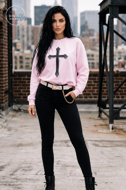 A woman stands on a rooftop wearing a light pink crewneck sweatshirt with a large black cross graphic on the front, paired with black skinny jeans, a white belt, and black boots. The background shows a city skyline and brick buildings.
