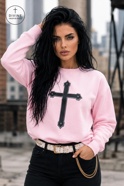 A woman wearing a light pink crewneck sweatshirt with a large black cross graphic on the front, paired with black jeans and a patterned belt. The background shows a blurred city rooftop setting.