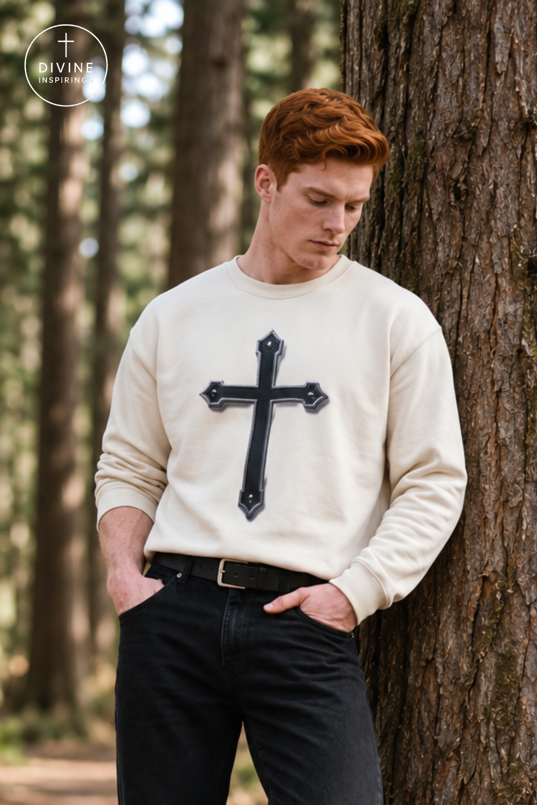 A man with red hair leans against a tree in a forest, wearing a cream-colored crewneck sweatshirt with a black cross graphic on the front, paired with black pants.
