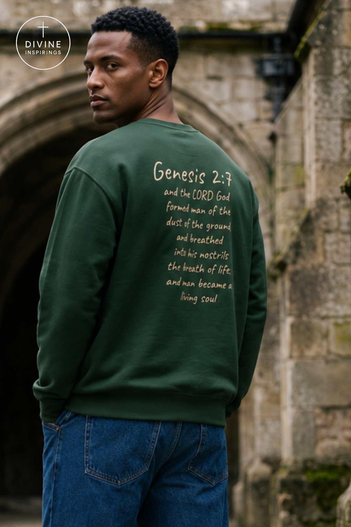 A man wearing a dark green crewneck sweatshirt with white printed scripture text on the back, standing outdoors in front of a stone archway. The Divine Inspirings logo is visible in the top left corner.