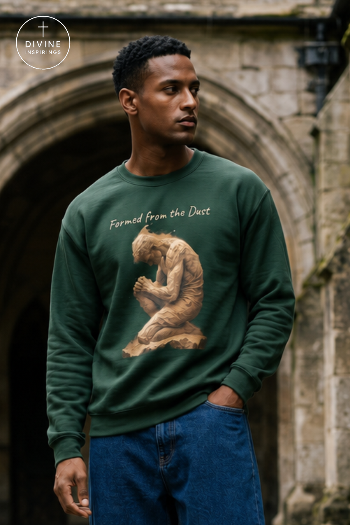 A man wearing a dark green crewneck sweatshirt with a graphic of a kneeling figure and the text 'Formed from the Dust', paired with blue jeans, standing in front of a stone archway. A 'Divine Inspirings' logo is visible in the top left corner.