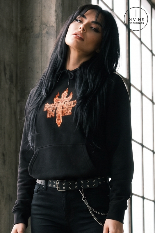 A woman with long black hair models a black hoodie featuring a graphic print of a flaming cross and the words 'REFINED IN FIRE' in orange and gold. She is wearing black jeans with a studded belt and a chain accessory.