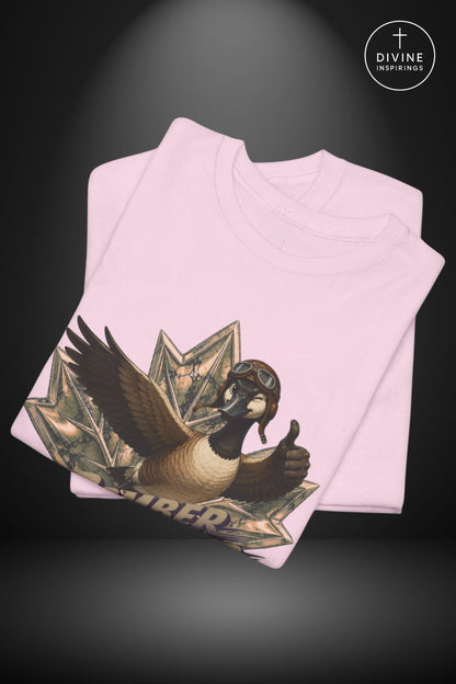 A folded light pink t-shirt featuring a graphic print of a goose wearing aviator goggles and giving a thumbs-up, emerging from a geometric star design. The shirt is displayed on a dark gray gradient background with a spotlight effect, and a 'Divine inspirings' logo is visible in the top right corner.