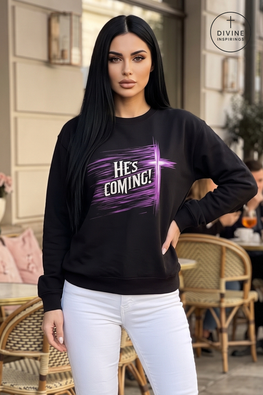 A woman modeling a black crewneck sweatshirt with a purple and white 'Hes Coming!' graphic, paired with white pants, set against an outdoor cafe background.