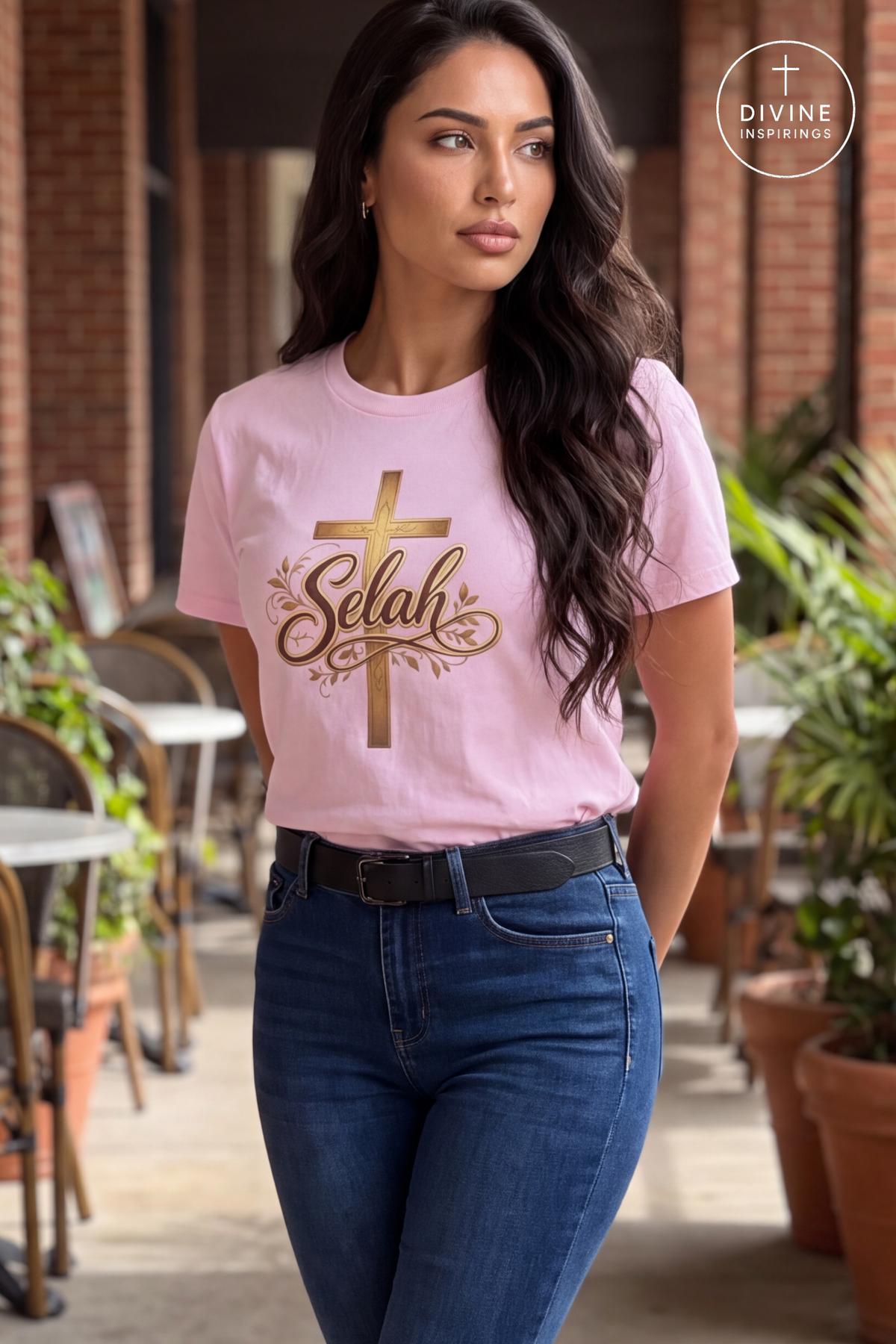 A woman wearing a light pink crewneck t-shirt with a gold cross and 'Selah' graphic, paired with dark blue jeans, standing in an outdoor patio setting.
