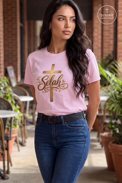 A woman wearing a light pink crewneck t-shirt with a gold cross and 'Selah' graphic, paired with dark blue jeans, standing in an outdoor patio setting.
