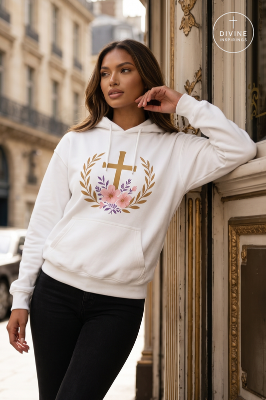 A woman wearing a white pullover hoodie with a gold cross, floral wreath, and lavender accents, posing outdoors against an ornate building facade.
