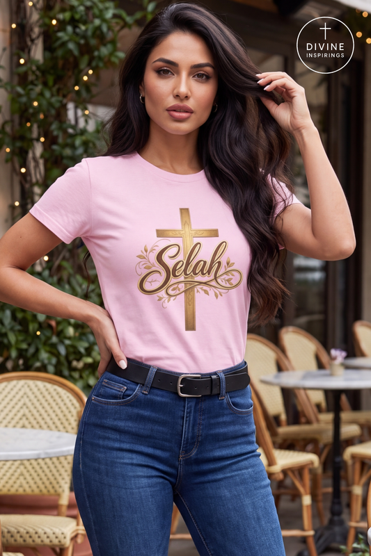 A woman wearing a light pink t-shirt with a gold cross and 'Selah' text graphic, paired with blue jeans, in an outdoor cafe setting. A 'DIVINE INSPIRINGS' logo is visible in the top right corner.