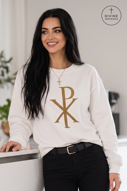 A woman wearing a white crewneck sweatshirt with a large gold Chi-Rho monogram on the front, paired with black pants and a gold necklace. The Divine Inspirings logo is visible in the top right corner.