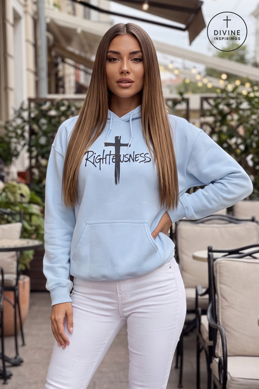 A woman modeling a light blue hoodie with a black cross and the word 'Righteousness' printed on the front, paired with white pants, set in an outdoor patio environment. A 'DIVINE INSPIRINGS' logo is visible in the top right corner.