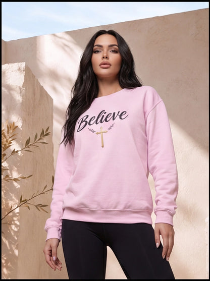 Woman wearing a pink sweatshirt with 'Believe' and a cross design against a beige wall.