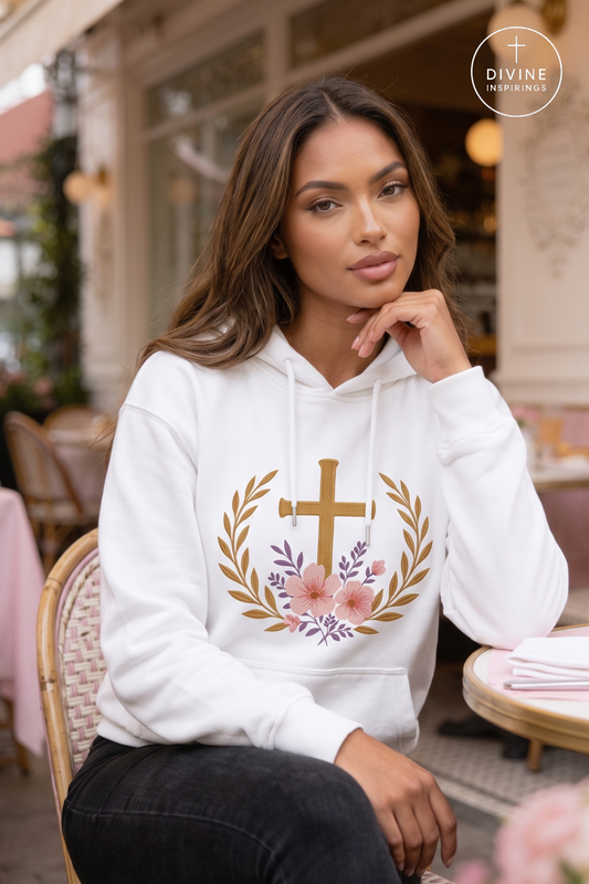 A woman wearing a white pullover hoodie with a gold cross, laurel wreath, and pink floral graphic on the front, seated at an outdoor cafe. A Divine Inspiration brand logo is visible in the top right corner.