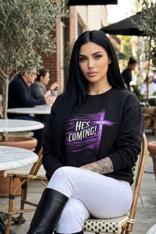 A woman with long black hair sits on a woven chair at an outdoor cafe, wearing a black crewneck sweatshirt with a purple graphic that reads 'He's COMING!' and white pants.