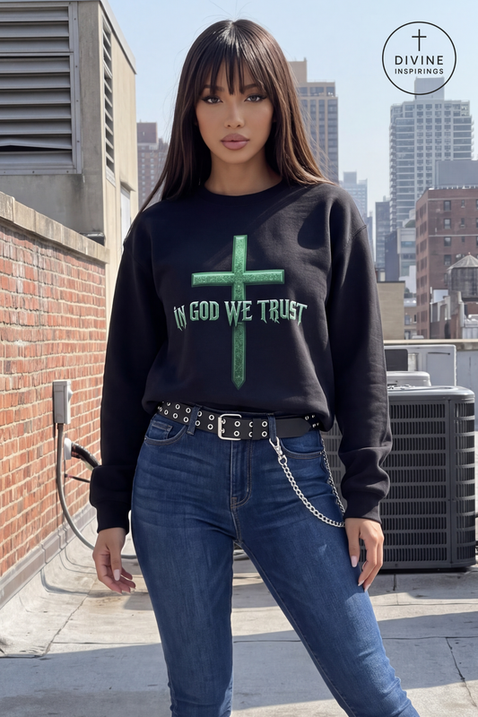 A woman stands on a city rooftop wearing a black crewneck sweatshirt with a green cross graphic and the words 'IN GOD WE TRUST' in green, paired with blue jeans and a black studded belt. A Divine Inspirings logo is visible in the top right corner.