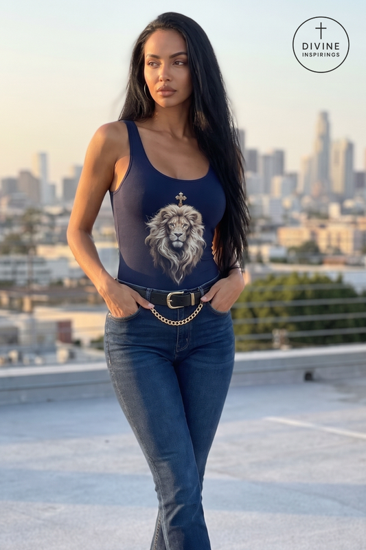 A woman stands on a rooftop wearing a navy blue tank top with a large lion graphic and a gold cross on the chest, paired with dark blue jeans and a gold chain belt. The background shows a blurred city skyline at sunset, and a 'DIVINE INSPIRINGS' logo is visible in the top right corner.