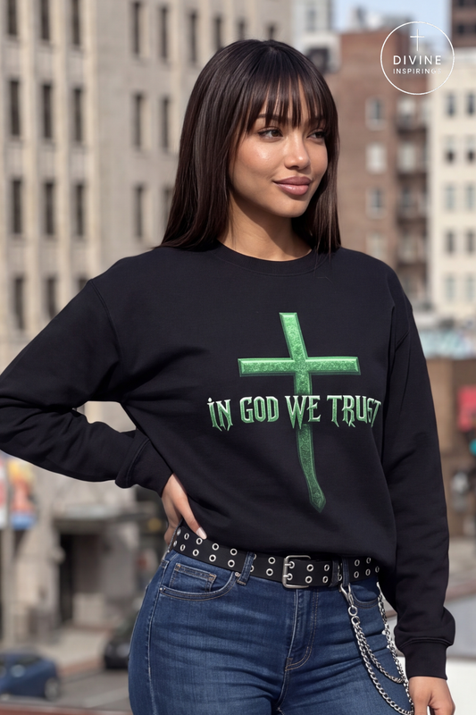A woman wearing a black crewneck sweatshirt with a green cross graphic and the text 'IN GOD WE TRUST' printed on the front, paired with blue jeans and a studded belt, set against a blurred city street background.