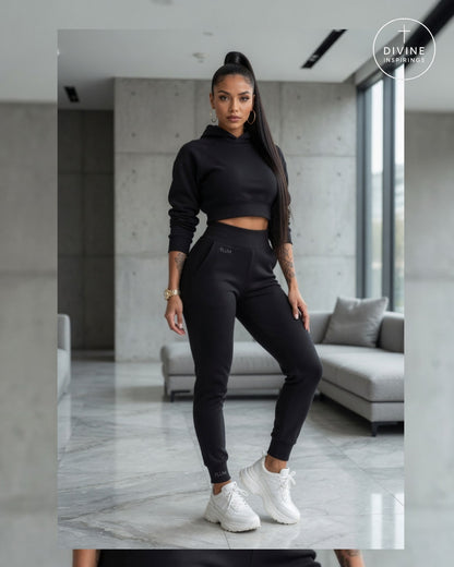 A woman modeling a black two-piece activewear set consisting of a cropped long-sleeve hoodie and matching jogger pants, paired with white sneakers, in a modern living room setting.